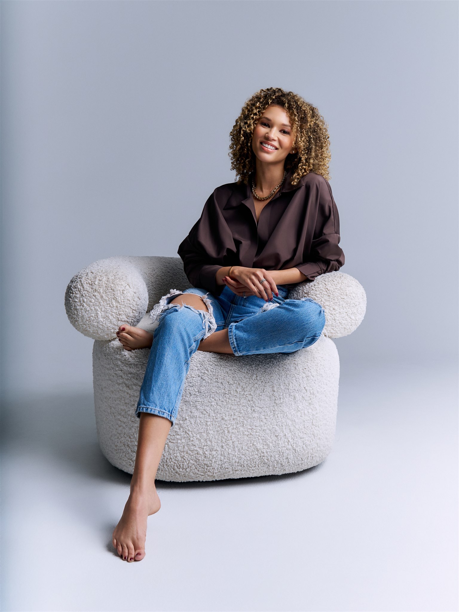 Samantha Neyland Trumbo in a brown v-neck shirt and ripped jeans sitting on a white bouncle chair.